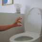 A hand holds The Unscented Company’s Toilet Bowl Cleaning Tab above an open toilet, ready to drop it in. A bag sits on a shelf and a framed picture hangs in the background, highlighting eco-friendly, sustainable hygiene.