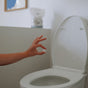 A hand holds The Unscented Company Toilet Bowl Cleaning Tab above an open toilet, ready to drop it in. In the background, a product packet and framed photo emphasize sustainable hygiene.