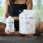 Dressed in black, a person sits cross-legged holding The Unscented Company’s Lotion Refill - Unscented in a pump bottle and pouch, both white with minimalist labels—ideal for moisturized skin.