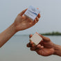 Two hands hold up a plastic free Shampoo Bar and its box, labeled "The Unscented Company," against a blurred background of water and trees under a cloudy sky.