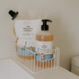 A bottle and refill pouch of The Unscented Company Kids Shiny Shampoo sit in a white basket on a bathroom shelf. Both feature playful, illustrated labels with children, emphasizing safe, natural ingredients.