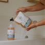A person refills a Kids Shiny Shampoo Refill Bag from The Unscented Company over a bathtub. Both packages feature playful illustrations, emphasizing natural ingredients and gentle care for the whole family.