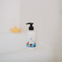 A white bottle of The Unscented Company Kids Smooth Conditioner with a black pump sits on a white bathtub edge, its child-friendly illustrated label visible; the hypoallergenic formula features naturally derived ingredients. A yellow rubber duck is blurred in the background.