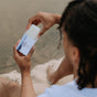A person with braided hair sits near water, holding an open white rectangular Soap Bar by The Unscented Company in their hands.