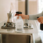 A person holds The Unscented Company’s All Purpose Cleaner - Unscented spray bottle by a kitchen sink, with a stovetop and moka pot visible in the background.