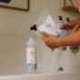 A person refills a bottle using The Unscented Company Kids Big Bubble Bath Refill Bag above a white bathtub with black faucets in the background.