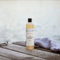 A bottle of The Unscented Company Body Soap sits on a wooden dock beside a folded striped towel, with tranquil water in the background.