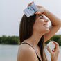 A woman smiles outdoors by the water, holding The Unscented Company’s Conditioner Bar in one hand, with greenery and sky in the background.