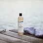 A bottle of The Unscented Company Daily Shampoo sits on a wooden dock next to a folded striped towel, with blurred water in the background.