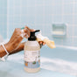 A person pumps Gentle Baby Wash + Shampoo by The Unscented Company from a bottle onto a yellow sponge in front of a blue tiled bathtub filled with bubbles, wearing a beaded bracelet and turquoise ring.
