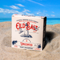 A box of Old Salt Hair/Beard Shampoo rests on a sandy beach with ocean and sky behind it. The packaging displays nautical designs and text noting it's suitable for both hair and beards.