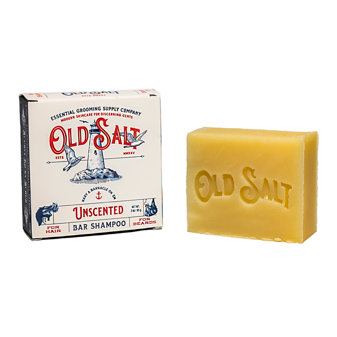 A rectangular bar of Old Salt Hair/Beard Shampoo, embossed with the brand name, sits beside its box featuring a lighthouse design and blue and red branding details. The unscented bar is ideal for hair and beard care.