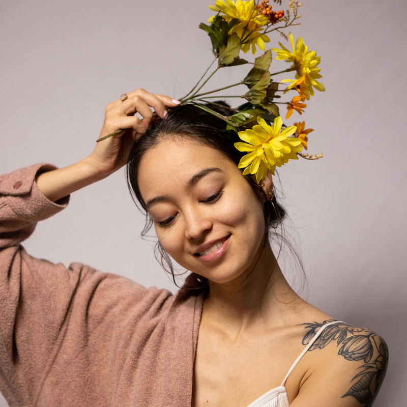 A woman with a floral tattoo on her shoulder smiles gently, holding yellow flowers above her head. She wears a light pink sweater draped over one shoulder and stands against a plain, soft background.