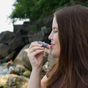 A woman with long brown hair and a nose piercing smiles with her eyes closed, holding a Chagrin Valley Soap & Salve Lip Balm Stick: Simply Natural Vegan near her lips while standing outside by rocks and greenery next to the water.