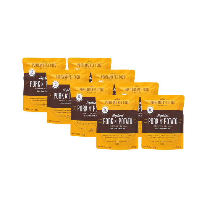 Six 9 oz BPA-free recyclable pouches of Portland Pet Food Companys Hopkins Pork N Potato Human-Grade Dog Meal, Mixer or Meal Topper, are arranged in a staggered row. The yellow and brown packaging features white text on a white background.