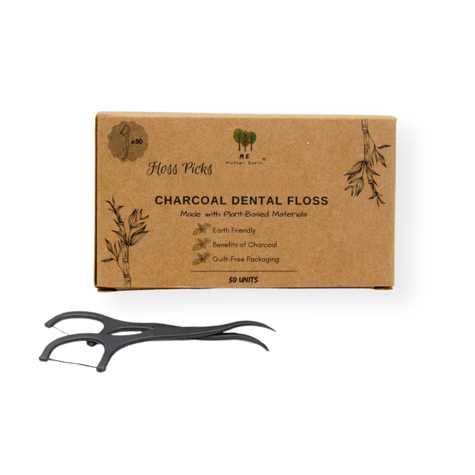 A brown box labeled "Me Mother Earth Charcoal Dental Floss Picks" with plant illustrations stands behind two black bamboo charcoal floss picks, featuring eco-friendly, plastic-free packaging and a cornstarch-based handle.