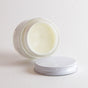 A glass jar of Little Seed Farm Booty Balm for Babies, an organic, cloth-diaper-safe balm made with healing herbs, lies on its side with the metal lid off and in front, set against a plain white background.