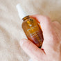 A hand holds a brown glass bottle of Little Seed Farm Beard Oil, a nourishing beard moisturizer, against a textured beige background.