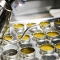 A metal kettle pours liquid into small, round tins of Little Seed Farm’s Hand-Aid Healing Hand Balm, ideal for dry, cracked hands. The neatly arranged tins suggest a professional production setting.