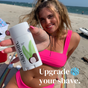 A smiling woman in a bright pink bikini holds out a travel-friendly Shavestix Solid Shave Stick on a sandy beach. The text reads, Upgrade your shave. The hydrating balm glows against the ocean and blue sky backdrop.