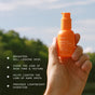 A hand holds an orange bottle of Ursa Major Brighten Up Vitamin C Serum outdoors, with trees and sky behind it. Text highlights its featherweight hydration and skin benefits, such as brightening and evening skin tone.