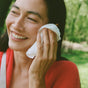 A woman in a red sweater with long dark hair smiles as she uses an individually wrapped Ursa Major Essential Face Wipe outdoors, surrounded by greenery.