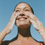 Beneath a clear blue sky, a smiling person cleanses their face with Ursa Major’s Fantastic Face Wash. Wet hair clings as gentle foam and sunlight reveal fresh, smooth skin on their face and hands.