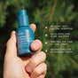 A hand holds a blue Ursa Major Forest Water Hyaluronic Serum bottle outdoors, with text showcasing its hydrating, plumping, and fast-absorbing benefits—perfect as a hyaluronic acid serum for dehydrated skin. Blurred green foliage is visible in the background.