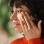 A curly-haired individual in an orange sweater gently touches their cheek, pondering the benefits of Ursa Majors Forest Water Hyaluronic Serum for dehydrated skin, against a softly blurred background highlighting their features.