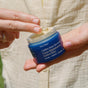 A person, dressed in a light-colored textured shirt, holds a blue jar labeled “Ursa Major Golden Hour Recovery Cream” in one hand and scoops out the hydrating face cream with the other.
