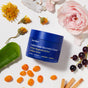 A blue jar of Ursa Major Golden Hour Recovery Cream sits among flowers, green leaves, blackcurrants, and orange berries on a white background, emphasizing its deep hydration benefits.