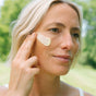 A woman with short blonde hair gently applies Ursa Major’s Golden Hour Recovery Cream to her cheek with her fingers while outdoors, treating her skin to deep hydration surrounded by lush, blurred greenery.