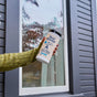 A person in a green sweater holds a container of Aunt Fannie's Glass + Window Cleaning Wipes in front of a window with gray siding.