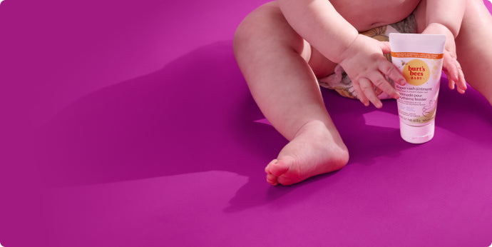 A baby sitting on a bright purple surface holds a tube of Burts Bees Baby ointment, with only the babys lower body visible.