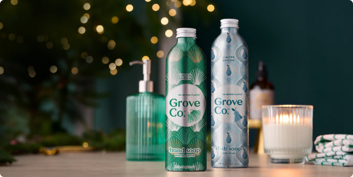 Two decorative aluminum bottles labeled Grove Co. stand on a table, one for hand soap and one for dish soap. They are surrounded by a candle, glass soap dispenser, and festive lights in the blurred background.