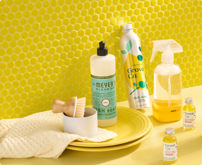 A yellow plate with a scrub brush, cleaning cloth, and various dish soaps and cleaners sits in front of a bright yellow, circular-tile backsplash.