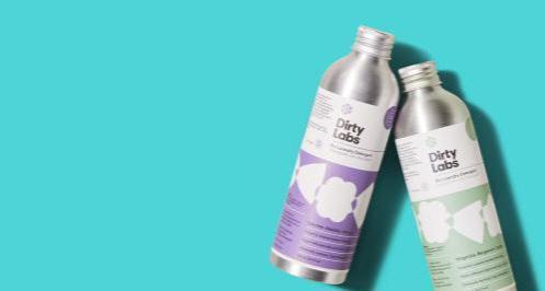Two metallic bottles of Dirty Labs detergent, one with a purple label and one with a green label, are placed on a turquoise background. The bottles are tilted and partially overlap.