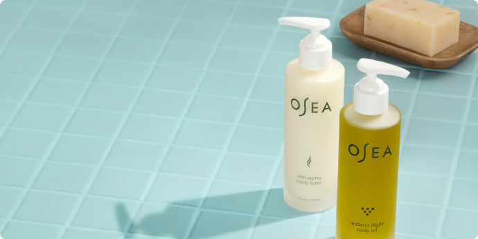 Two OSEA bottles, one white and one green, with pump tops sit on a light blue tiled surface next to a beige bar of soap on a wooden dish.