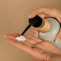 A close-up of hands dispensing Pacha Soap Co. Body Lotion with hydrating coconut oil from a pump bottle onto the palm, set against a neutral beige background.