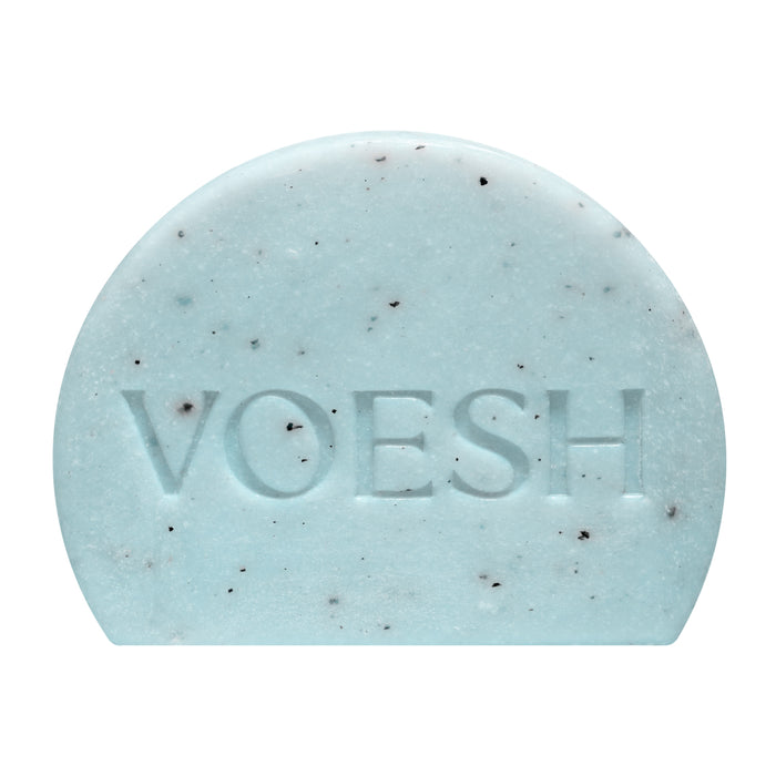 The VOESH Body Buffing Bar With AHA is a light blue, half-circle exfoliator with dark specks and “VOESH” engraved on the front.