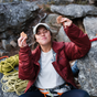 A smiling person in a maroon jacket and cap sits outdoors on rocks, holding up two Kates Real Food Organic Energy Bars - White Chocolate Macadamia. Climbing gear nearby suggests theyre taking a break during a rock climbing adventure.