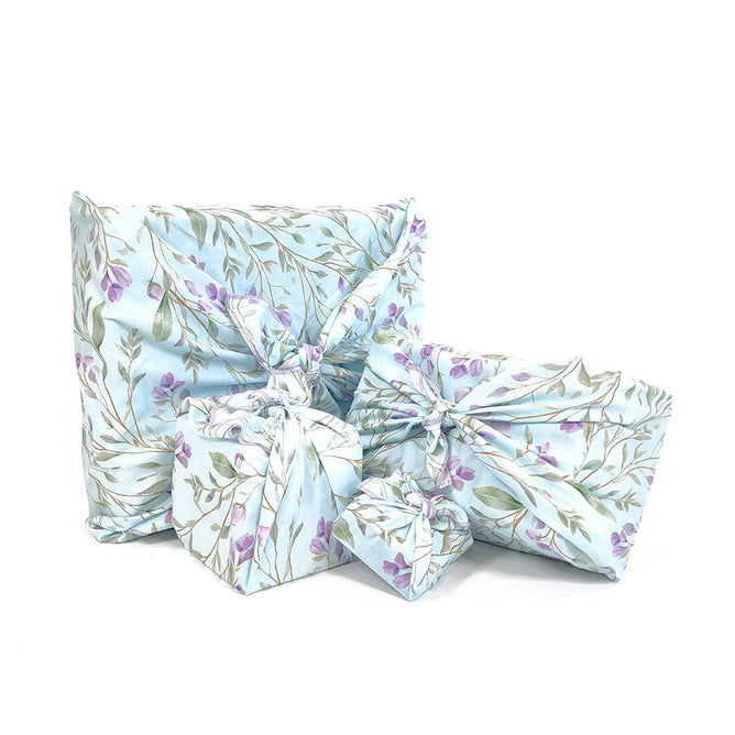 Three gifts wrapped using Waste Free Celebrations Furoshiki feature a floral pattern with green leaves and purple flowers on light blue fabric. Tied with a knot, they showcase an eco-friendly technique, offering a sustainable alternative to traditional wrapping methods.