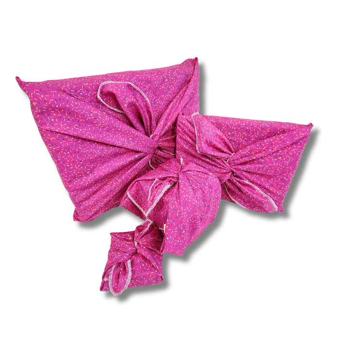 Four Furoshiki gift boxes from Waste Free Celebrations, wrapped in vibrant pink fabric with white and purple speckles, are arranged on a white background. Each box features a bow made from reusable fabric wraps—an elegant and eco-friendly alternative to conventional wrapping.