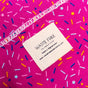 Close-up of vibrant pink Furoshiki fabric with colorful sprinkle patterns. A white label reads Waste Free Celebrations, Made in Afghanistan with a website link. This sustainable option features stitched white threading, ideal for reusable gift wraps.