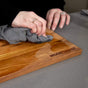A person cleans a wooden cutting board engraved with "WOOD FOR CHEF" using a gray cloth. The board is treated with Food Grade Mineral Oil for Wood Cutting Board by WoodForChef.
