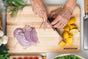 A person slices a red onion and lemons on the Mizu Minimalist Chopping Board by WoodForChef, crafted from 100% FSC-certified maple wood, surrounded by fresh vegetables and kitchen tools.