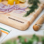 The Mizu Minimalist Chopping Board by WoodForChef, made from 100% FSC-certified maple wood, holds sliced red onions, lemon wedges, and fresh herbs. Cherry tomatoes and a towel are nearby, with a wooden-handled knife on the counter.