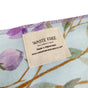 A close-up of Waste Free Celebrations Furoshiki, featuring a floral fabric wrap with purple flowers and green leaves on a light background. The tag reads Made in Afghanistan www.wastefreecelebrations.com, showcasing sustainable gift wrapping for eco-friendly festivities.