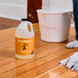 Aunt Fannie's Hardwood Floor Cleaner, Value Size, made with essential oils, sits on a wooden floor beside a white bucket, mop, and someone in white socks.
