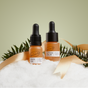 The UpCircle Winter Glow Duo, featuring two amber dropper bottles, stands in snow with green foliage and a gold ribbon—an ideal sustainable gift set against a light green background.
